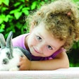 little beautiful girl smiling, hugging a baby rabbit on the green background in summer. happy laughing child and pet playing outdoors. female looking in the camera. bunny is a symbol of Easter.