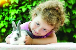 little beautiful girl smiling, hugging a baby rabbit on the green background in summer. happy laughing child and pet playing outdoors. female looking in the camera. bunny is a symbol of Easter.