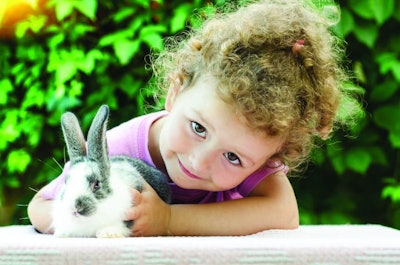 little beautiful girl smiling, hugging a baby rabbit on the green background in summer. happy laughing child and pet playing outdoors. female looking in the camera. bunny is a symbol of Easter.