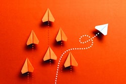 White paper plane is flying to the right on orange colored background where a group of orange colored paper planes are flying straight up.