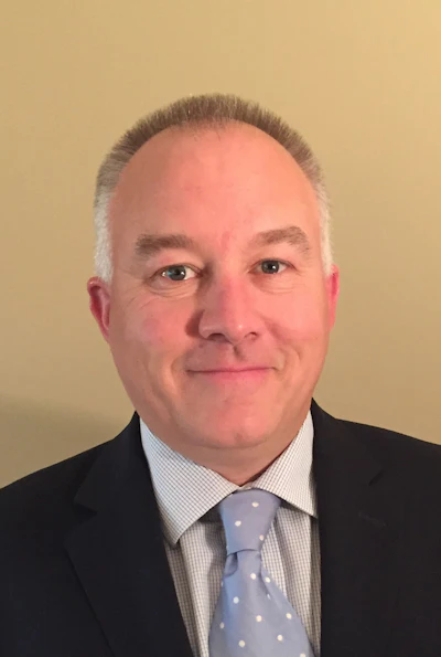 Professional headshot of a man in dark suit, light blue checkered shirt, and patterned tie against neutral background