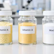 Three glass jars labeled Vitamin K, Vitamin A, and Niacin containing supplement powders in a laboratory setting