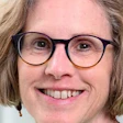 Professional headshot of a smiling woman wearing black-framed glasses with shoulder-length brown hair