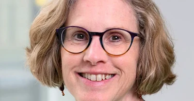 Professional headshot of a smiling woman wearing black-framed glasses with shoulder-length brown hair