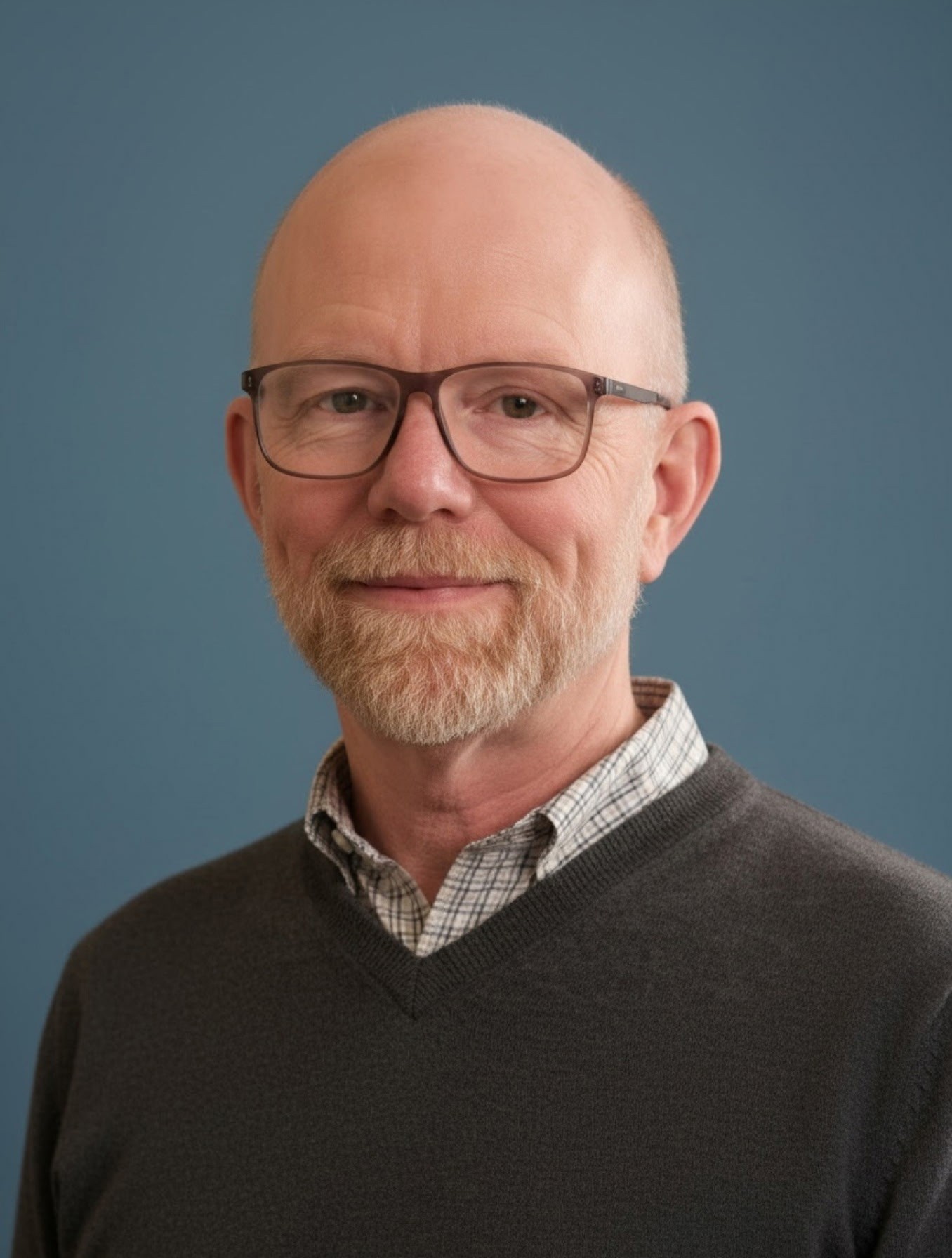Professional headshot of a bearded man wearing glasses and a dark sweater over a checkered shirt against a blue background