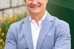 Professional headshot of a smiling man in light blue blazer and white shirt against green background with yellow flowers