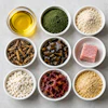 Overhead view of cooking ingredients in bowls including oil, green powder, flour, nuts, capers, pink salt, white cheese, beets, and oats on gray surface