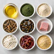 Overhead view of cooking ingredients in bowls including oil, green powder, flour, nuts, capers, pink salt, white cheese, beets, and oats on gray surface