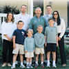 Multi-generational family of nine posing together outdoors in front of a white house with green landscaping