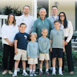 Multi-generational family of nine posing together outdoors in front of a white house with green landscaping