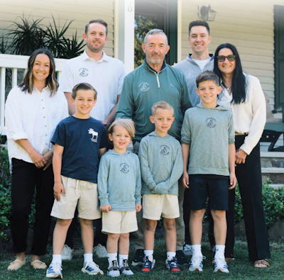 Multi-generational family of nine posing together outdoors in front of a white house with green landscaping