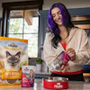 Woman with purple hair holding Nulo cat food product in modern kitchen with pet food packaging and red bowl