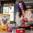 Woman with purple hair holding Nulo cat food product in modern kitchen with pet food packaging and red bowl