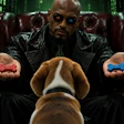 Man in black leather coat and sunglasses holds red and blue pills while seated, with brown dog in foreground and digital code background