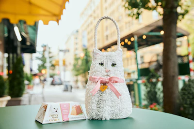 White plush cat-shaped gift bag with pink bow and gold heart charm on outdoor café table