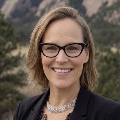 Professional headshot of a smiling woman wearing glasses and dark blazer against a mountainous backdrop