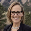 Professional headshot of a smiling woman wearing glasses and dark blazer against a mountainous backdrop