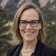 Professional headshot of a smiling woman wearing glasses and dark blazer against a mountainous backdrop