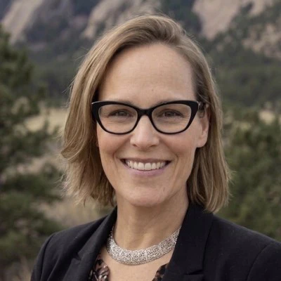 Professional headshot of a smiling woman wearing glasses and dark blazer against a mountainous backdrop