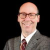 Professional headshot of a smiling man wearing glasses, gray plaid blazer, and burgundy tie against black background