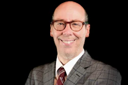 Professional headshot of a smiling man wearing glasses, gray plaid blazer, and burgundy tie against black background