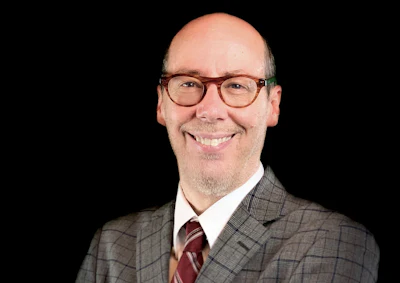 Professional headshot of a smiling man wearing glasses, gray plaid blazer, and burgundy tie against black background