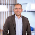 Professional man smiling in modern office environment wearing brown blazer and light blue shirt