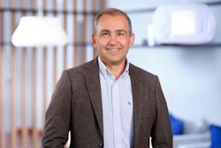 Professional man smiling in modern office environment wearing brown blazer and light blue shirt