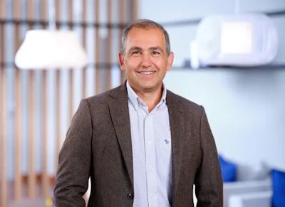 Professional man smiling in modern office environment wearing brown blazer and light blue shirt