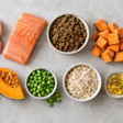 Healthy pet food ingredients including salmon, chicken, blueberries, sweet potato, carrots, oats, and supplements arranged on gray background