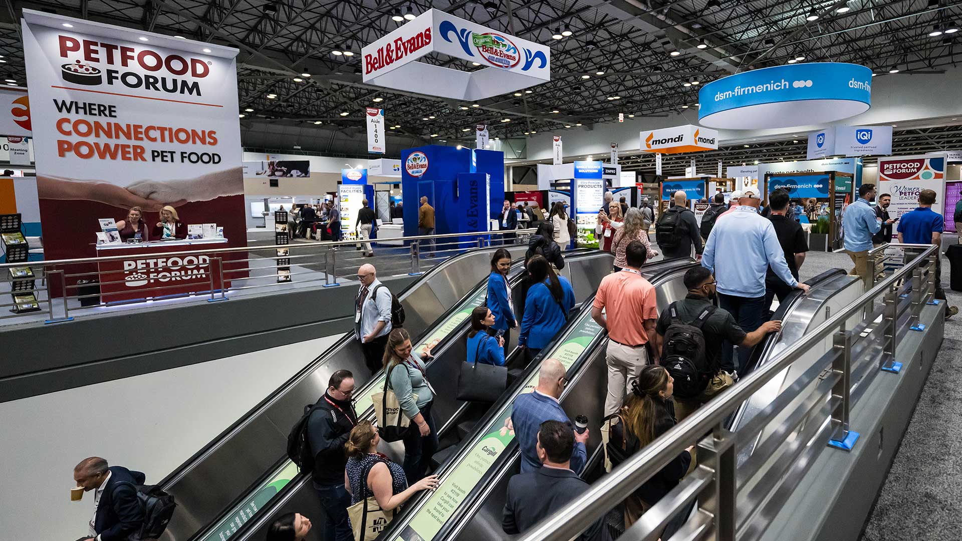 Crowded pet food industry trade show floor with escalators, vendor booths, and branded signage including Petfood Forum banner.