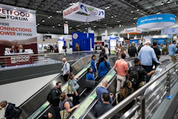 Crowded pet food industry trade show floor with escalators, vendor booths, and branded signage including Petfood Forum banner.
