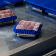 Packaged bacon or cured meat in blue hinged containers on a stainless steel surface in a commercial kitchen or retail setting