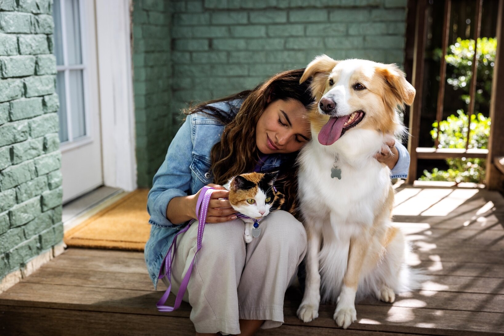 Woman smiling while holding a cat and posing with a golden and white dog on a sunny patio