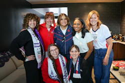 Seven women smiling together at an indoor event, some wearing USA team apparel and medals