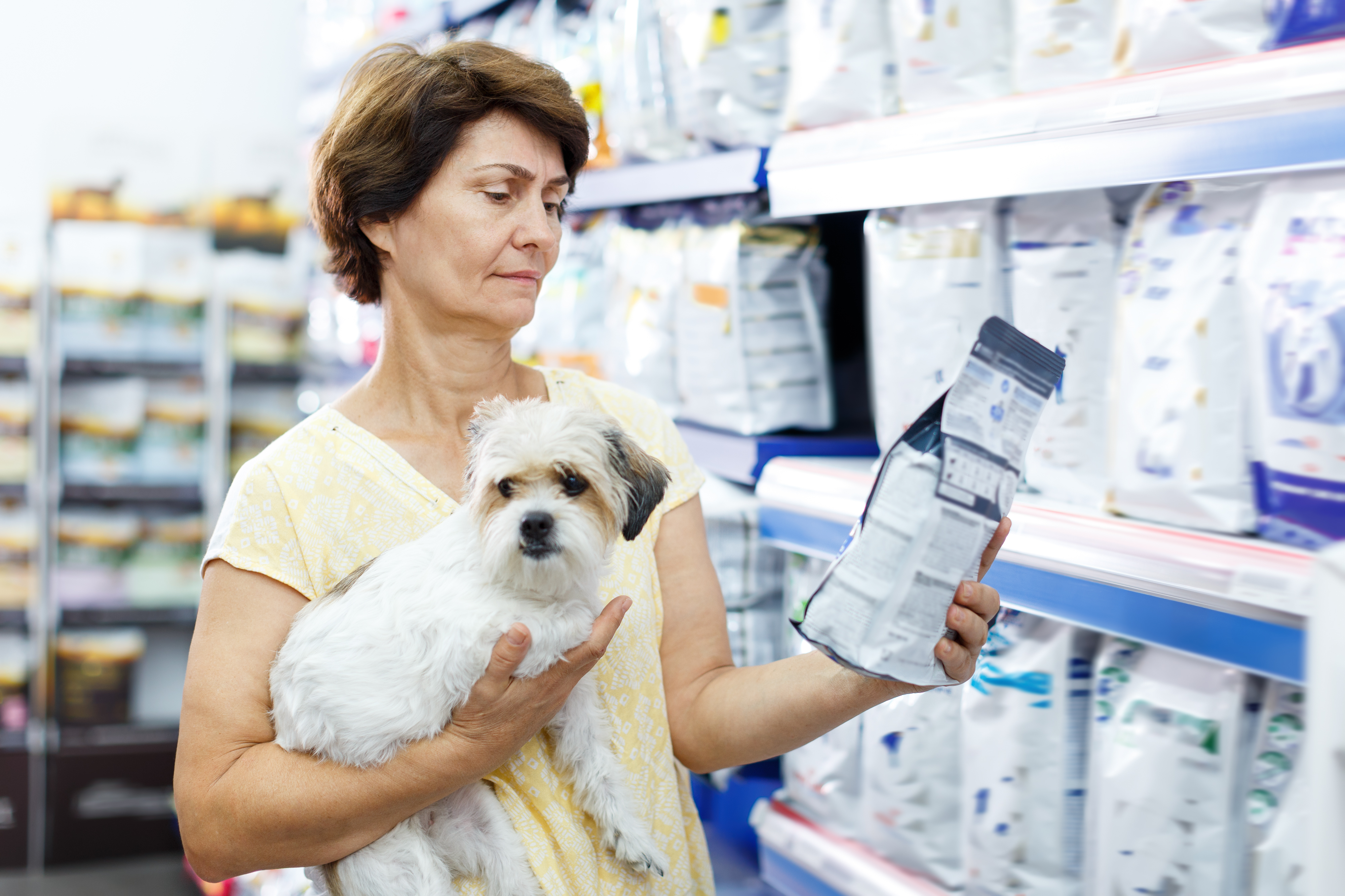 Woman holding small white dog while reading medication or pet product package in pharmacy aisle
