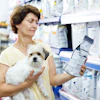 Woman holding small white dog while reading medication or pet product package in pharmacy aisle