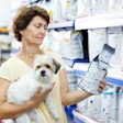 Woman holding small white dog while reading medication or pet product package in pharmacy aisle