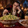 Aristocratic man in period costume dining alone in candlelit Gothic hall with wine and grapes