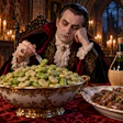 Aristocratic man in period costume dining alone in candlelit Gothic hall with wine and grapes
