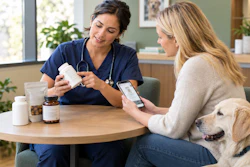 Healthcare provider in blue scrubs discussing medication with patient holding smartphone, accompanied by golden retriever